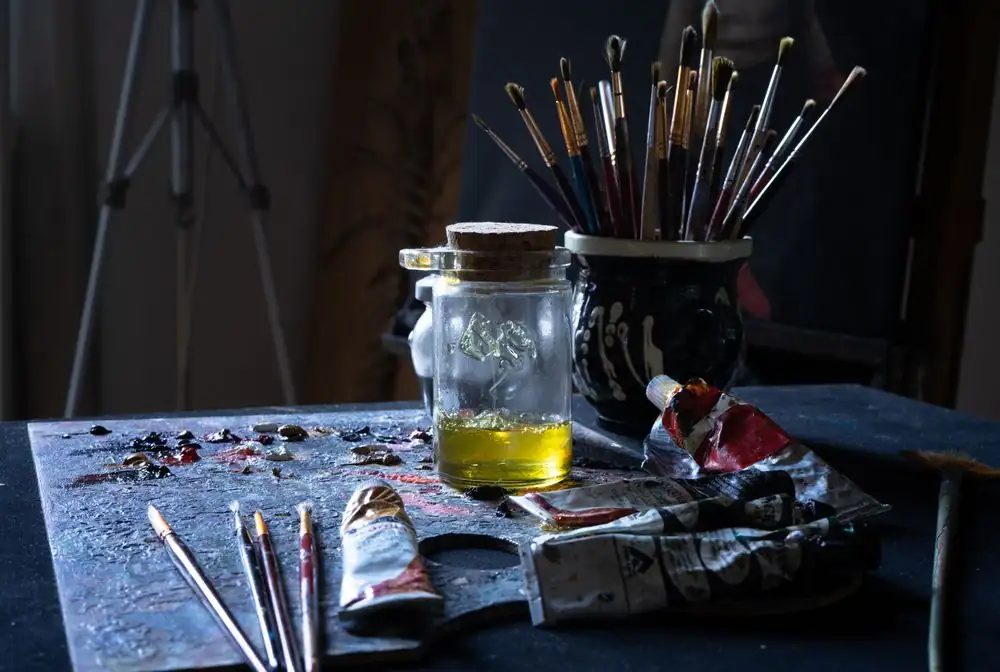 A cluttered artist's table filled with paint tubes, brushes, and a glass jar.