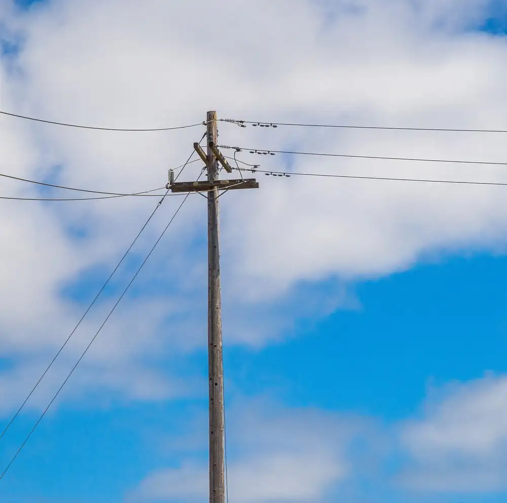 Wooden power pole with transmission lines against a clear blue sky and clouds.