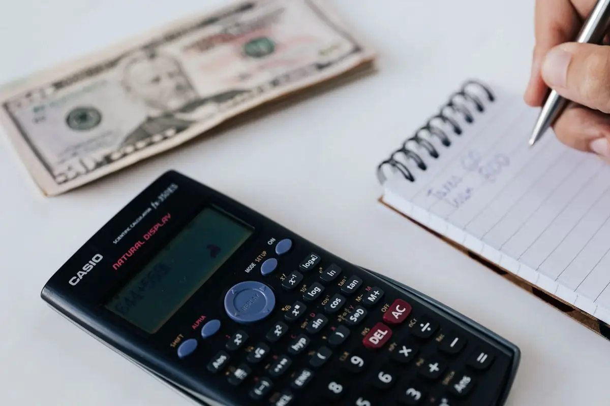 Close-up of a calculator, US dollar bill, and a hand writing in a notebook, symbolizing financial planning.