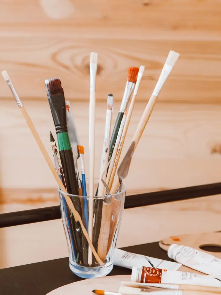Vertical shot of various paintbrushes in a glass holder on an artist's workspace.